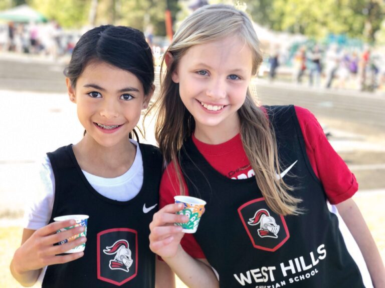 Two elementary girls in jerseys smile for the camera near track and field.
